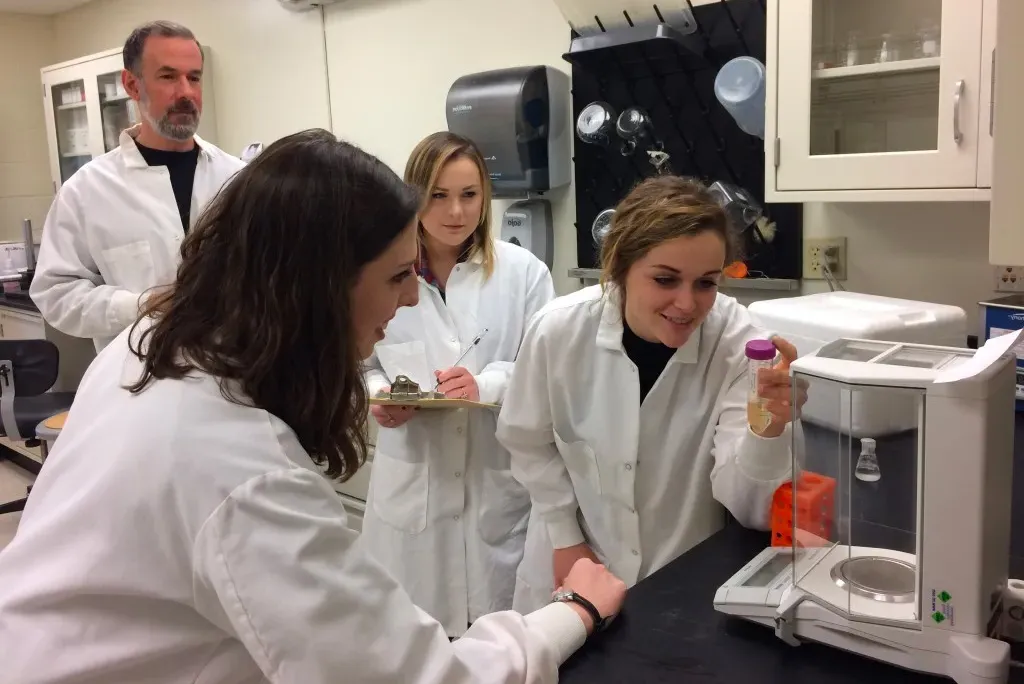 Three students and a professor wearing white coats and working in a lab setting