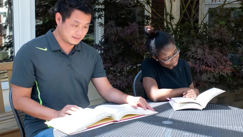 Two U N E students sit together at an outside table each reading a textbook